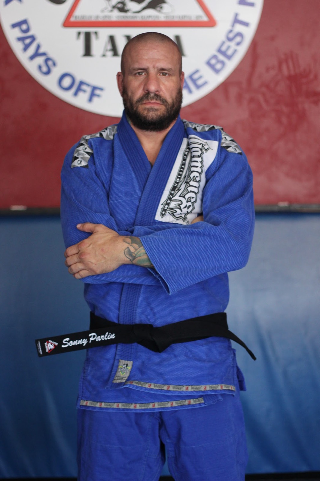 Sonny Parlin standing in a blue gi with arms crossed, wearing a black belt embroidered with his name